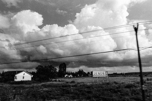 Black-and-white landscape photograph of a rural roadside scene with expansive storm clouds, power lines crossing the frame, a small house, and a roadside billboard reading “JESUS,” captured in an open prairie setting.